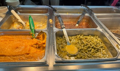 A food serving station with trays of orange mashed sweet potatoes, green beans, and a brown dish, all with serving utensils.