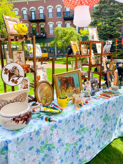 A vibrant outdoor display of vintage items, including framed art, ceramics, and colorful decorations on a floral tablecloth.
