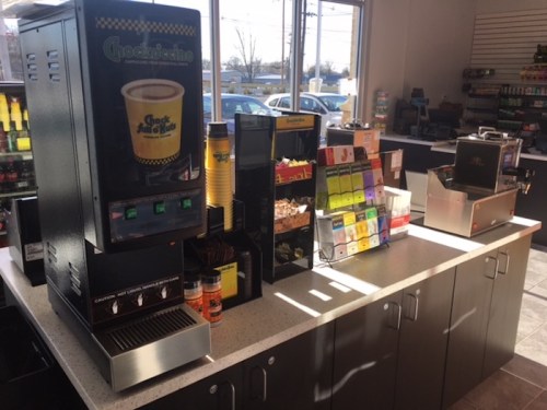 A coffee station with a machine, cups, and various snacks displayed in a bright convenience store.