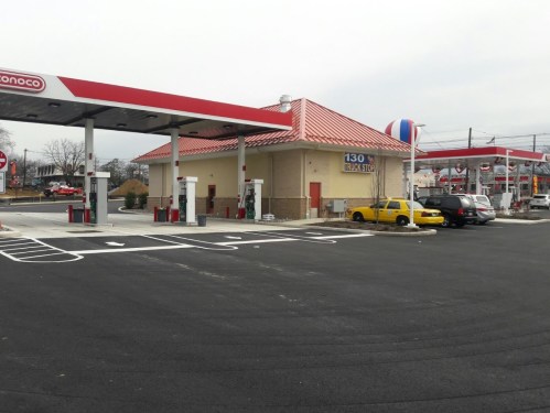 A gas station with a red and white roof, featuring fuel pumps and a yellow taxi parked nearby.