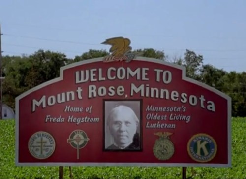 Welcome sign for Mount Rose, Minnesota, featuring a portrait of Freda Hegstrom and local symbols.