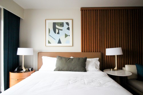 A cozy hotel room featuring a bed with a green pillow, modern artwork, and stylish lamps beside a side table.