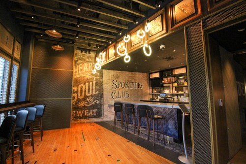 Interior of a stylish bar named "The Sporting Club," featuring wooden floors, high stools, and vintage decor.