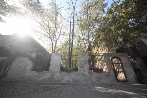 A sunlit scene of a textured wall with a gate, surrounded by trees and plants, creating a serene outdoor atmosphere.