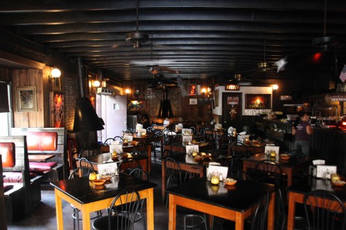 Cozy restaurant interior with wooden beams, dark tables, and warm lighting, featuring empty booths and a bar area.