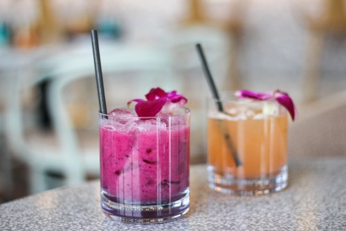 Two colorful cocktails with straws, one pink with flowers and one orange, served in clear glasses on a textured surface.