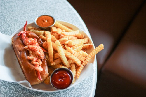 A plate featuring a lobster roll with fries and two dipping sauces on a textured table.