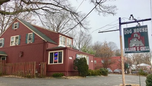A red building with green shutters, featuring a sign that reads "Dog House Saloon & Grill" in a wooded area.