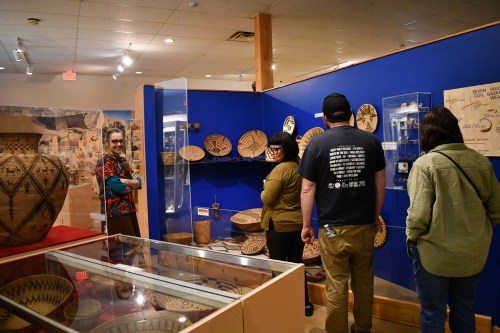 Visitors admire Native American baskets and pottery in a museum exhibit, with vibrant blue walls and display cases.