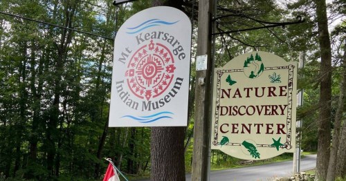 Two signs on a tree: one for Mt. Kearsarge Indian Museum and another for Nature Discovery Center, surrounded by greenery.
