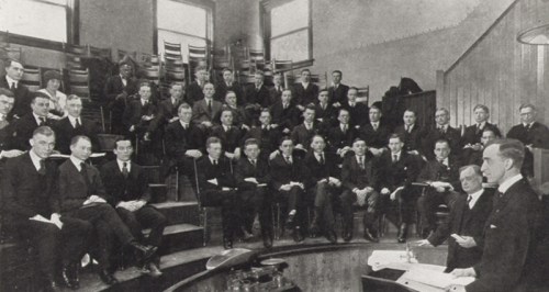 A historical group photo of men in formal attire seated in a lecture hall, attentively listening to a speaker.