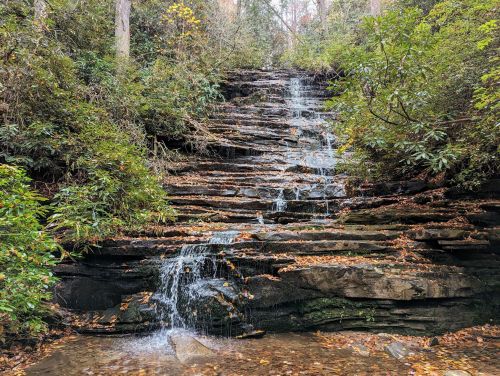 A serene waterfall cascading over rocky steps, surrounded by lush greenery and autumn leaves.