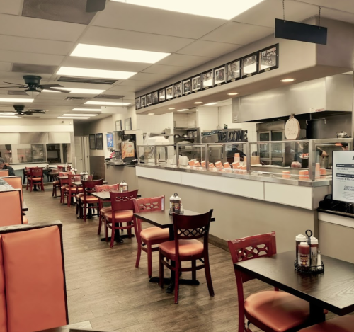 Interior of a casual restaurant featuring orange booths, wooden tables, and a counter with a kitchen in the background.