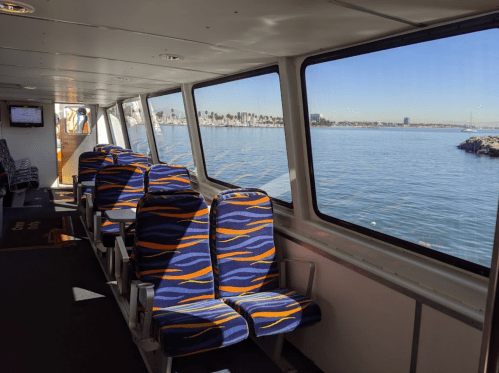 Interior of a ferry with colorful seats, large windows showing a calm water view and a clear blue sky.