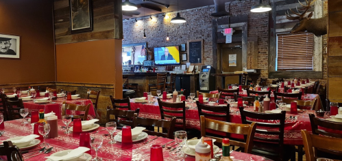 Interior of a restaurant with wooden decor, red tablecloths, and tables set for dining. A TV is visible in the background.