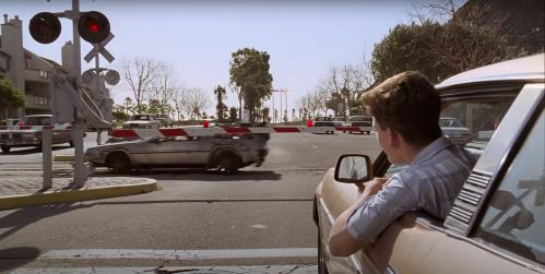A person leans out of a car window as a train passes by at a crossing, with palm trees in the background.