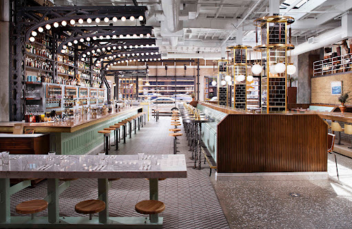 A stylish bar interior featuring a long counter, high stools, and shelves lined with bottles under bright lights.
