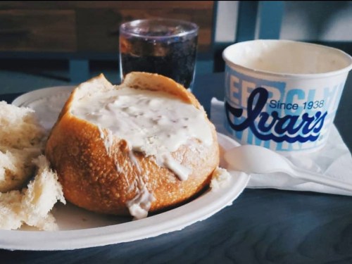 A bread bowl filled with clam chowder, accompanied by a drink and a piece of bread on a plate.