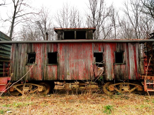 An old, weathered train caboose with missing windows, surrounded by bare trees and overgrown grass.