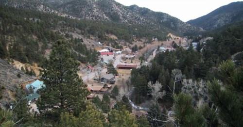 Aerial view of a small mountain town surrounded by trees and hills, with buildings scattered throughout the valley.