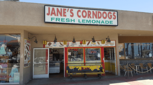 Exterior of Jane's Corndogs shop, featuring a bright sign and a colorful entrance with a menu display.