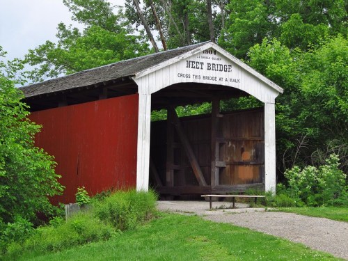 A red-covered bridge with a white roof, surrounded by greenery, inviting visitors to cross at a walk.