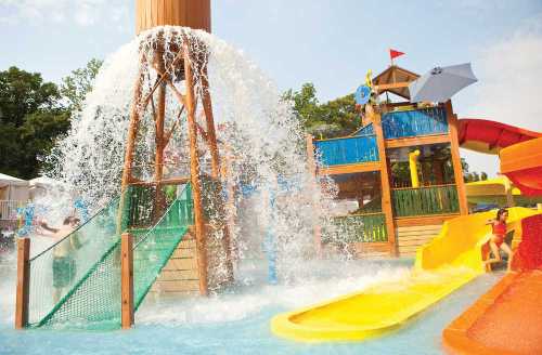 A colorful water park with slides and a large water tower splashing down water, surrounded by trees and blue sky.