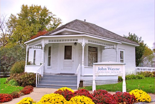 Historic white house with a porch, surrounded by colorful flowers, featuring a sign that reads "John Wayne."