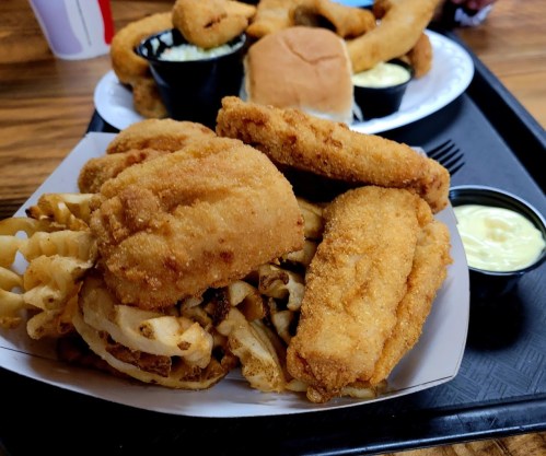 A plate of crispy fried chicken pieces on a bed of waffle fries, with a side of dipping sauce and a bun.