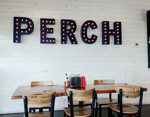A cozy dining area with a wooden table and chairs, featuring a large illuminated sign that reads "PERCH."
