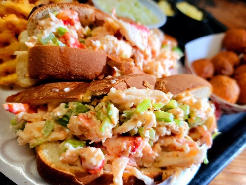 A close-up of a lobster roll with chopped lobster and celery on toasted bread, served with waffle fries and hushpuppies.