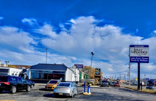 A busy parking lot in front of Jolly Roger's restaurant, with blue skies and scattered clouds above.