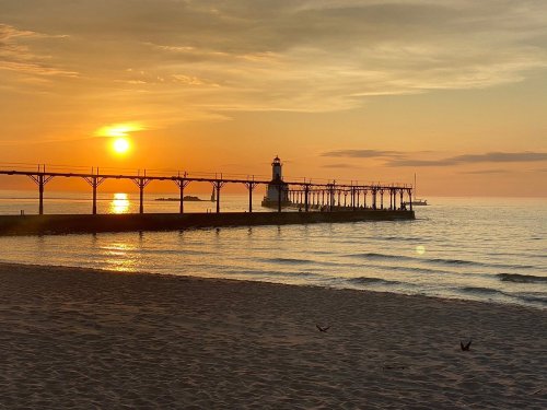 A serene sunset over a calm lake, featuring a lighthouse on a pier and gentle waves lapping at the sandy shore.