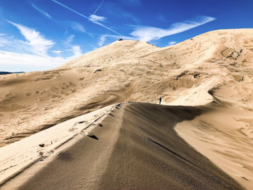 A lone figure walks along a sandy dune under a blue sky with wispy clouds.