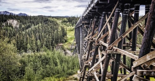 A wooden trestle bridge spans a lush green landscape with trees and hills in the background under a cloudy sky.