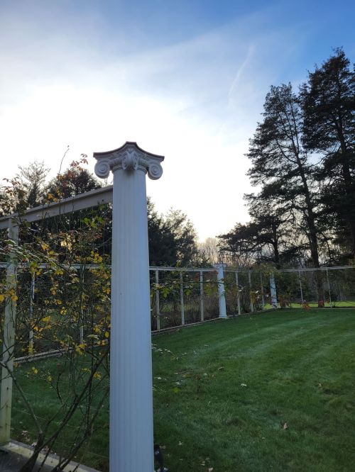 A white column stands in a garden, with trees and a clear sky in the background.