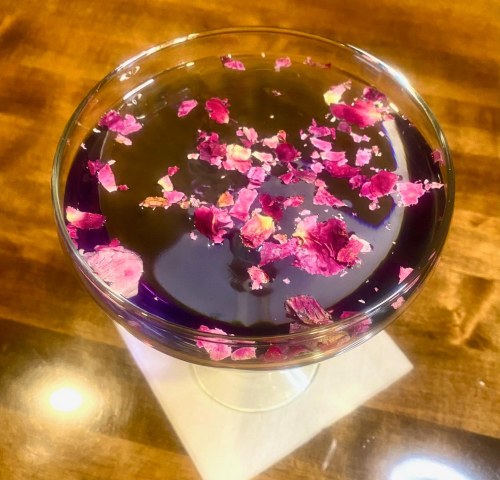 A cocktail in a glass, topped with colorful rose petals, resting on a napkin on a wooden table.