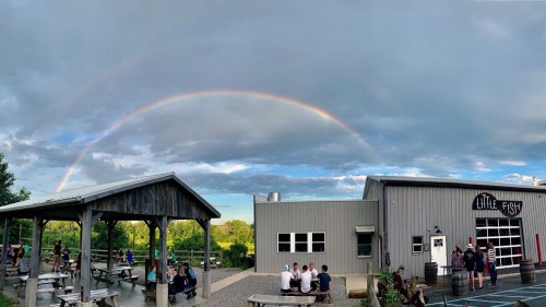 A brewery with a rainbow overhead, featuring outdoor seating and people enjoying the space under a cloudy sky.