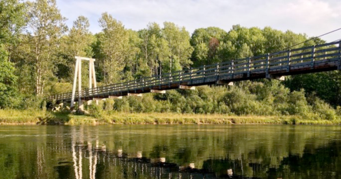 A wooden suspension bridge spans a calm river, surrounded by lush green trees and a clear blue sky.
