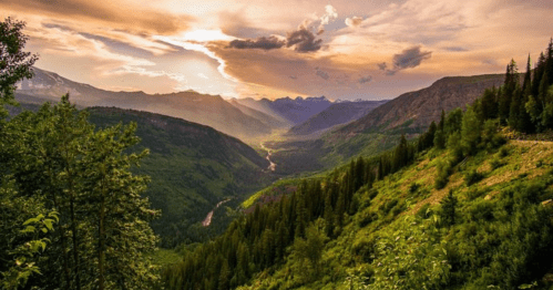 A scenic view of a valley surrounded by mountains, with a river winding through lush greenery under a colorful sunset.
