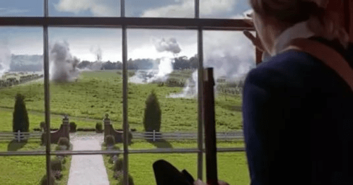 A person looks out a window at a battlefield with smoke rising from explosions in the distance.