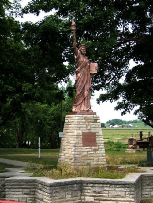 A bronze statue resembling the Statue of Liberty stands on a stone pedestal, surrounded by greenery.
