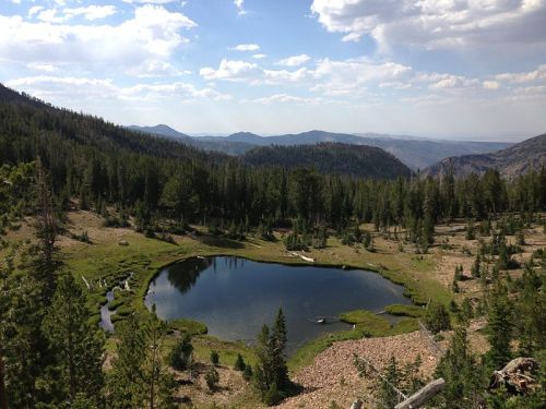 A serene landscape featuring a small pond surrounded by lush greenery and mountains under a partly cloudy sky.