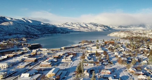 Aerial view of a snowy town by a lake, surrounded by mountains under a clear blue sky.