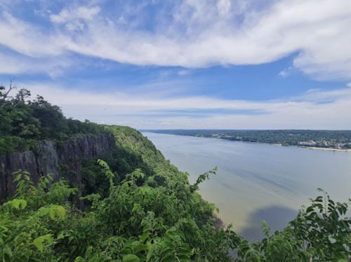 A scenic view of a river surrounded by lush greenery and cliffs under a blue sky with scattered clouds.