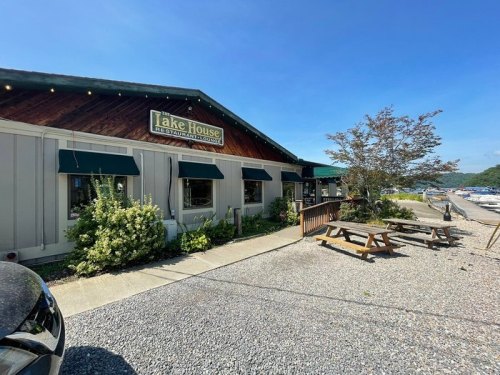A lakeside restaurant with a wooden exterior, green awnings, and outdoor seating, surrounded by trees and a clear blue sky.