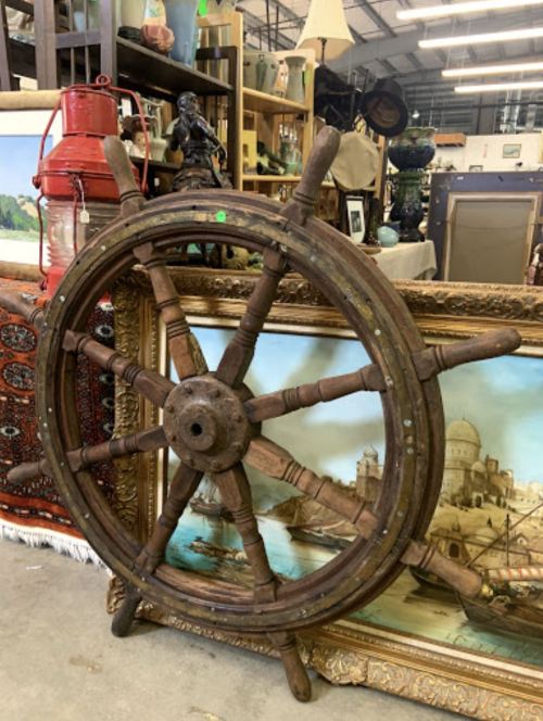 A vintage wooden ship's wheel displayed in an antique shop, surrounded by various decorative items and paintings.