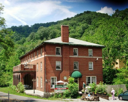 A brick inn surrounded by greenery, featuring a porch and a sign welcoming guests.