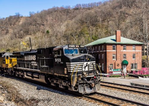 A black train engine passes by a brick building in a rural area, surrounded by trees and hills.