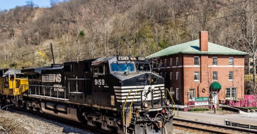 A black and white freight train passes by a brick building in a hilly, wooded area on a sunny day.